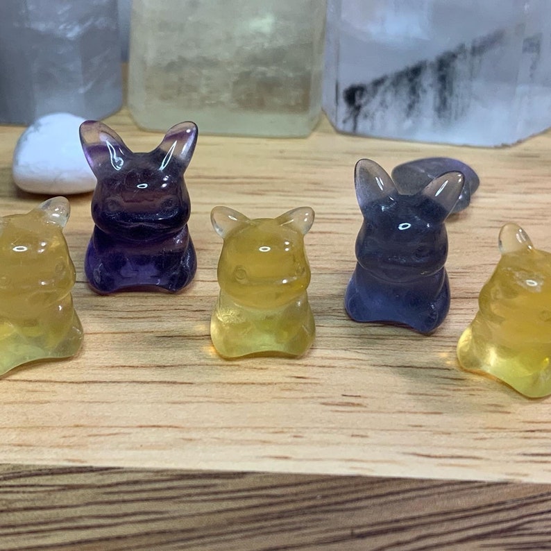 May include: Five small, hand-carved crystal figurines in the shape of a cartoon character. Two are yellow and three are purple.