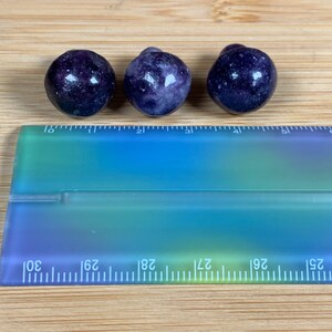 K&ouml;nnte beinhalten: Drei runde, dunkelviolette Edelsteine mit gesprenkelter Textur sind auf einer Holzoberfl&auml;che angeordnet. Ein transparenter Lineal mit blau-gr&uuml;nen Farbverl&auml;ufen befindet sich darunter, mit Messungen in Zentimetern.