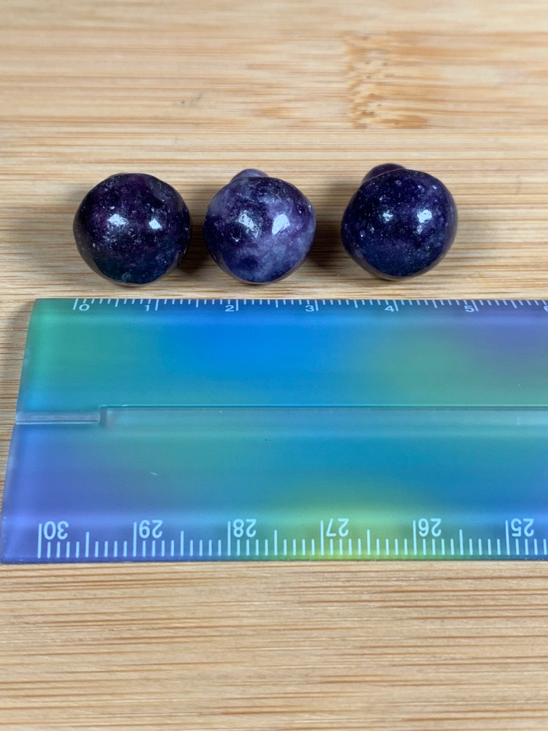 K&ouml;nnte beinhalten: Drei runde, dunkelviolette Edelsteine mit gesprenkelter Textur sind auf einer Holzoberfl&auml;che angeordnet. Ein transparenter Lineal mit blau-gr&uuml;nen Farbverl&auml;ufen befindet sich darunter, mit Messungen in Zentimetern.