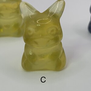 May include: A translucent yellow figurine of a cartoon character with large ears and a wide smile. The figurine is sitting with its legs crossed and its arms folded. The figurine is labeled with the letter "C".