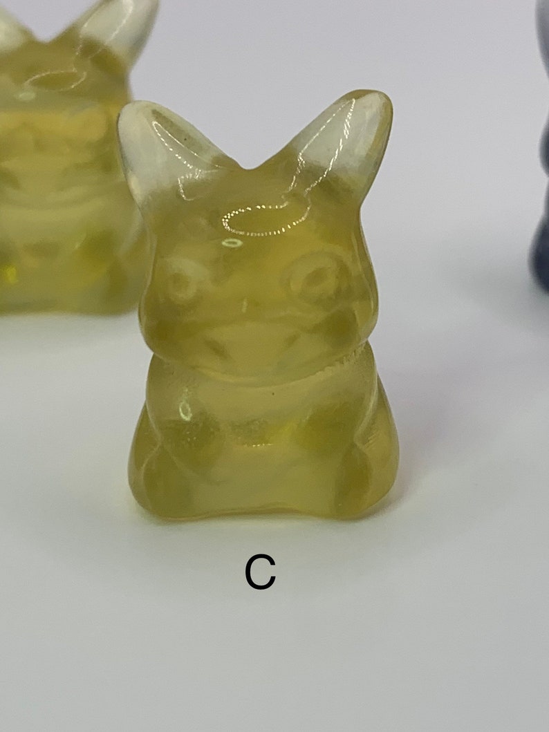 May include: A translucent yellow figurine of a cartoon character with large ears and a wide smile. The figurine is sitting with its legs crossed and its arms folded. The figurine is labeled with the letter "C".