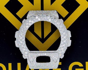 May include: A silver-colored watch case, encrusted with numerous small, sparkling stones. The watch case has a rounded shape with a large central opening and a rectangular section at the bottom. The background is black with gold lettering and a gold logo.