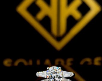 Può includere: Un anello di fidanzamento in argento con una pietra centrale rotonda e trasparente e due pietre più piccole e trasparenti su entrambi i lati. L'anello è su uno sfondo nero.