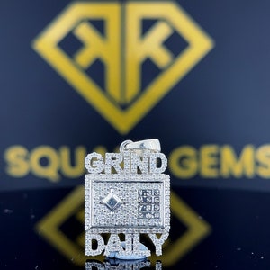 Op de afbeelding: Zilveren hanger met de woorden "GRIND DAILY" bezet met diamanten. De hanger heeft een klein vierkant met een diamant in het midden en een numeriek toetsenbord.