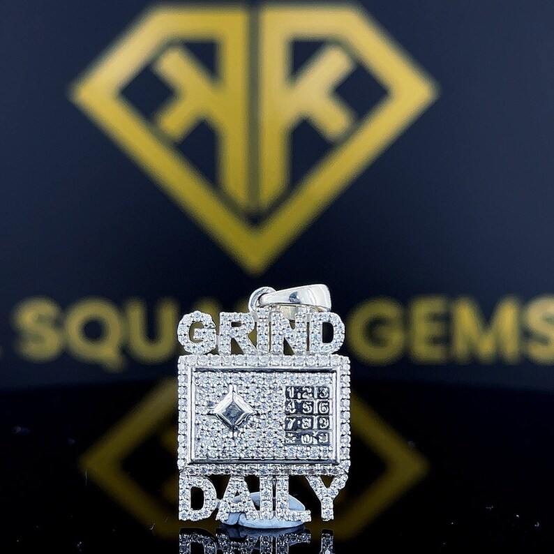Op de afbeelding: Een zilveren hanger met de woorden "GRIND DAILY" bezet met heldere zirkonia-stenen. De hanger heeft een klein vierkant met een diamantvormige steen in het midden en een rooster met nummers.