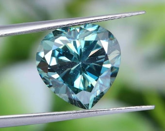 May include: A heart-shaped, blue moissanite gemstone with a brilliant cut. The gemstone is held by tweezers against a green background.