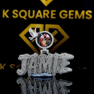 May include: A silver pendant spelling out "JAMIE" in a sparkling, diamond-like font. The pendant features a circular photo of a person and a dove. The background is black with gold lettering.