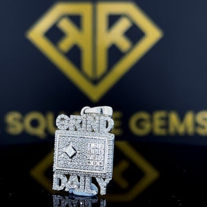 Op de afbeelding: Een zilveren hanger met een ontwerp dat is bezet met diamanten. De hanger heeft de woorden "GRIND DAILY" en een met diamanten bezet vierkant met de nummers 1 tot en met 9 erin.