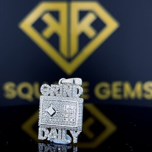 Op de afbeelding: Zilveren hanger met de woorden "Grind Daily" bezet met diamanten. De hanger heeft een klein diamantje in het midden van een vierkant met de nummers 1 tot en met 9.