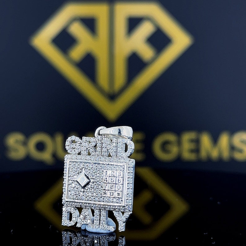Op de afbeelding: Zilveren hanger met de woorden "Grind Daily" bezet met diamanten. De hanger heeft een klein diamantje in het midden van een vierkant met de nummers 1 tot en met 9.