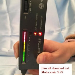 May include: A black diamond tester with a green and red light display. The tester is labeled "cultt" and "SELECTOR II". The text "LAMP ON BATTERY OK" and "LAMP ON READY OK" are also visible. The tester is being used to test a diamond. The text "Pass all diamond test Mohs scale: 9.25" is visible below the tester.
