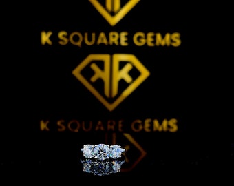 Può includere: Un anello di fidanzamento a tre pietre con un diamante rotondo brillante centrale e due diamanti rotondi brillanti più piccoli su entrambi i lati. L'anello è incastonato su una fascia in oro bianco ed è esposto su una superficie nera. L'anello si riflette sulla superficie.