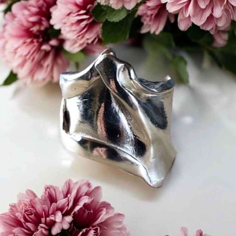 May include: A silver-colored, abstract cuff bracelet with a rippled, flowing design. The bracelet is set against a backdrop of pink flowers, creating a contrast between the metallic sheen and the soft floral hues. The cuff's form suggests a draped fabric.