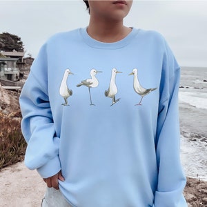 May include: A light blue sweatshirt featuring a whimsical illustration of four seagulls in various poses. The birds are depicted in shades of gray, white, and brown, with thin black legs and beaks. The sweatshirt is a soft, comfortable material.