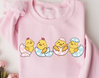 Peut inclure: Un sweat-shirt rose avec un motif de quatre poussins jaunes éclosant d'œufs. Les poussins sourient et ont les ailes déployées. Les œufs sont décorés de cœurs roses et bleus.