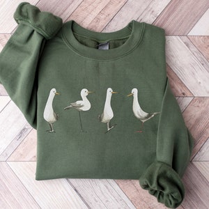 May include: An olive green sweatshirt featuring a watercolor illustration of four seagulls in various poses. The birds are depicted in shades of gray and white, with long, slender legs and beaks. The sweatshirt has a crew neck and long sleeves.