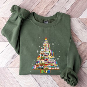 May include: A green sweatshirt with a Christmas tree design made of stacked books. The tree is decorated with a red star on top and snowflakes.