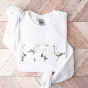 May include: White sweatshirt featuring a watercolor illustration of four seagulls in various poses. The birds have gray wings and are standing on thin black legs. The sweatshirt is made of a soft material and is suitable for casual wear.