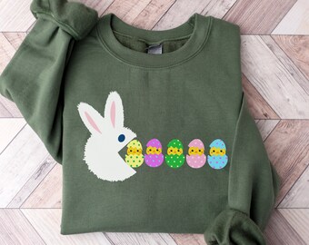 Peut inclure: Un sweat-shirt vert avec un lapin blanc qui mange une rangée d'œufs de Pâques colorés avec des poussins à l'intérieur. Les œufs sont décorés de pois et de rayures.