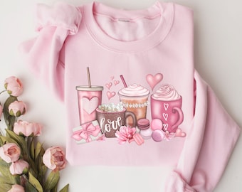 Puede incluir: Una sudadera rosa claro con un gráfico de varias bebidas rosas, que incluyen una taza para llevar, un café y una taza con crema batida. La palabra "love" está en la taza. También se presentan corazones rosas y macarons.