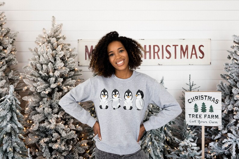 Puede incluir: Una mujer lleva una sudadera gris con un dise&ntilde;o de cuatro ping&uuml;inos en la parte delantera. La sudadera tiene cuello redondo y mangas largas. La mujer est&aacute; de pie frente a una pared blanca con dos &aacute;rboles de Navidad cubiertos de nieve a cada lado de ella. Hay un letrero encima de ella que dice "Feliz Navidad" y un letrero frente a ella que dice "Granja de &aacute;rboles de Navidad Corta el tuyo."