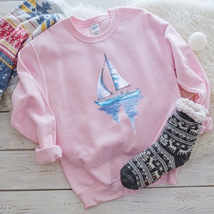 Pode incluir: Uma camisola rosa com um design de veleiro de aquarela. O veleiro &eacute; azul e branco com um reflexo na &aacute;gua. A camisola est&aacute; sobre uma superf&iacute;cie de madeira branca com um cobertor branco felpudo e um par de meias com padr&atilde;o preto e branco.