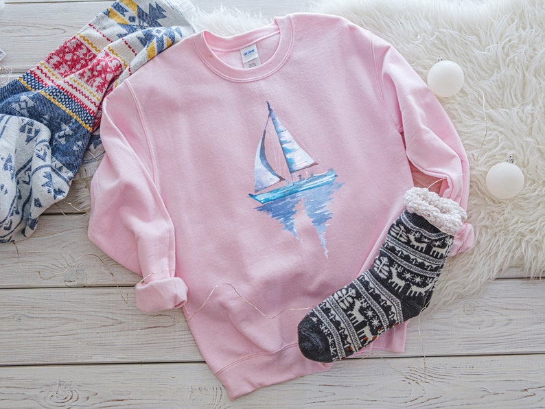Pode incluir: Uma camisola rosa com um design de veleiro de aquarela. O veleiro &eacute; azul e branco com um reflexo na &aacute;gua. A camisola est&aacute; sobre uma superf&iacute;cie de madeira branca com um cobertor branco felpudo e um par de meias com padr&atilde;o preto e branco.