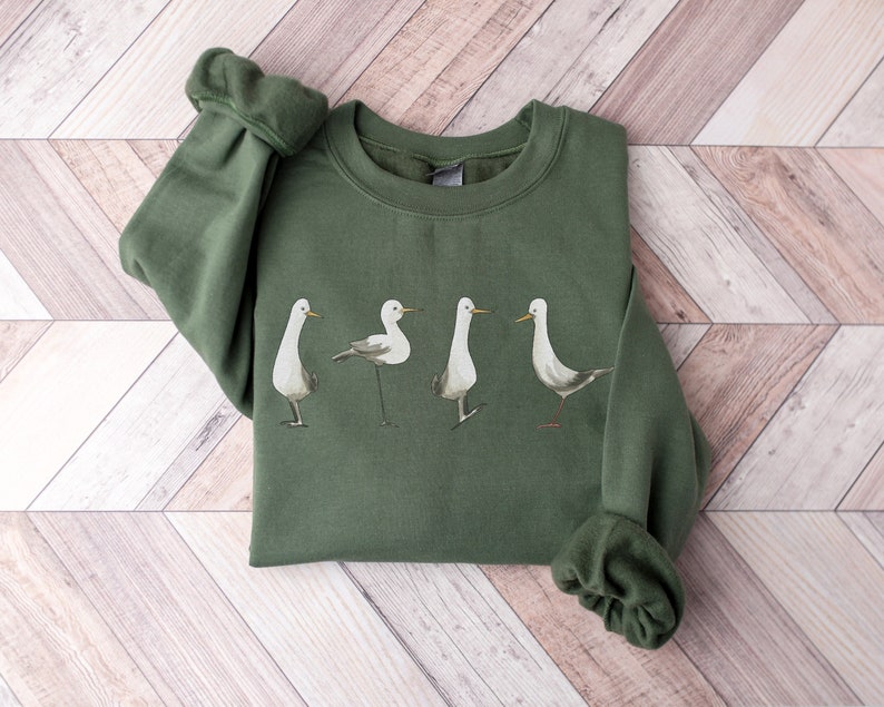 May include: An olive green sweatshirt featuring a watercolor illustration of four seagulls in various poses. The birds are depicted in shades of gray and white, with long, slender legs and beaks. The sweatshirt has a crew neck and long sleeves.