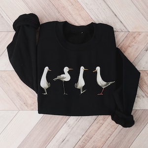 May include: Black sweatshirt featuring a watercolor design of four stylized birds in various poses. The birds are primarily white and gray with yellow beaks and red legs, set against a solid black background. The sweatshirt has a crew neck and long sleeves.