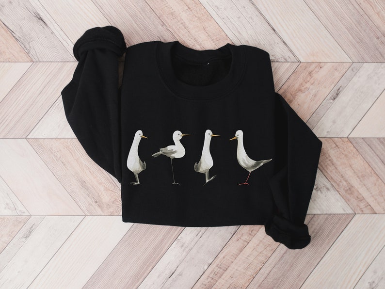 May include: Black sweatshirt featuring a watercolor design of four stylized birds in various poses. The birds are primarily white and gray with yellow beaks and red legs, set against a solid black background. The sweatshirt has a crew neck and long sleeves.