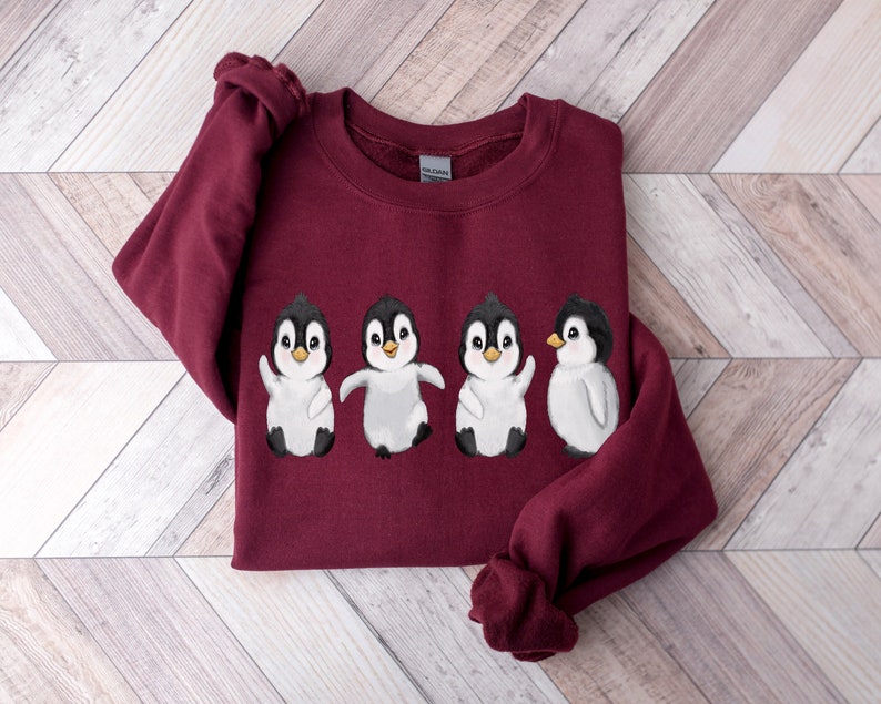 Puede incluir: Una sudadera color burdeos con un gr&aacute;fico de cuatro ping&uuml;inos en fila. Los ping&uuml;inos son blancos con ojos, picos y patas negras. Los ping&uuml;inos est&aacute;n todos mirando hacia adelante y tienen diferentes expresiones.