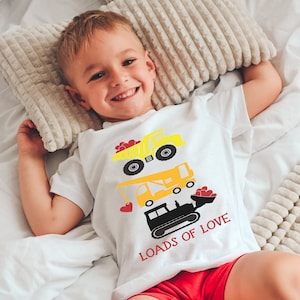 May include: A white t-shirt with a yellow construction truck, crane, and bulldozer graphic. The graphic has red hearts and the text "Loads of Love". A young child is wearing the shirt and lying on a white bed.