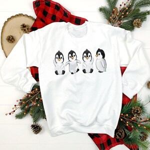 Puede incluir: Sudadera blanca con un gr&aacute;fico de cuatro ping&uuml;inos en fila. Los ping&uuml;inos est&aacute;n todos mirando hacia adelante y tienen las alas extendidas. Los ping&uuml;inos son negros y blancos con picos naranjas.