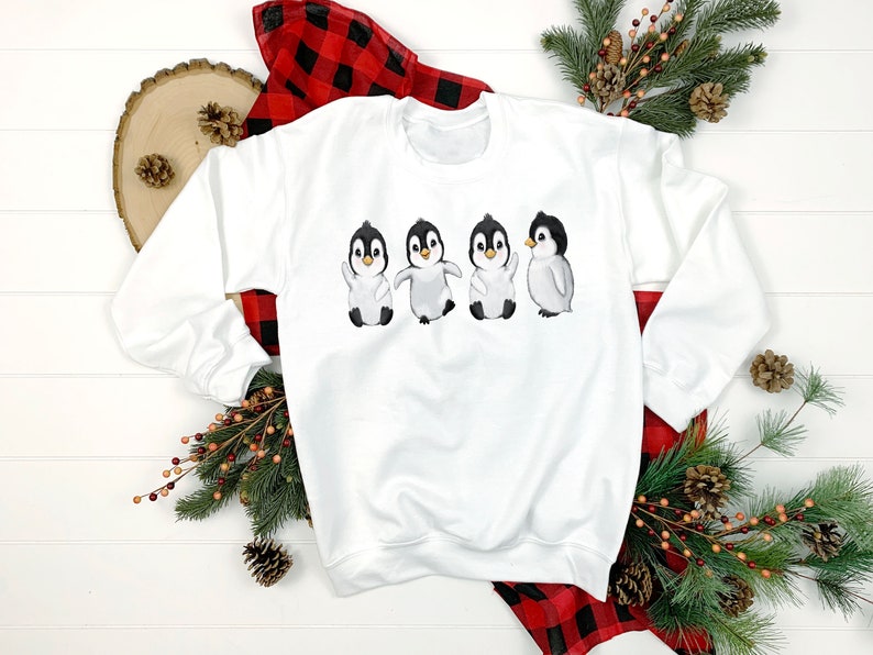 Puede incluir: Sudadera blanca con un gr&aacute;fico de cuatro ping&uuml;inos en fila. Los ping&uuml;inos est&aacute;n todos mirando hacia adelante y tienen las alas extendidas. Los ping&uuml;inos son negros y blancos con picos naranjas.