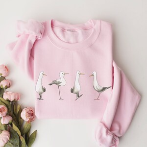 May include: A light pink sweatshirt featuring a watercolor illustration of four seagulls in various poses. The birds are depicted in shades of gray and white with orange beaks, adding a touch of whimsy to this casual apparel.