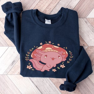 Peut inclure: Un sweat-shirt bleu marine avec un mignon dessin anim&eacute; d'axolotl portant un chapeau de champignon rouge. L'axolotl est entour&eacute; de feuilles vertes et de fleurs roses.