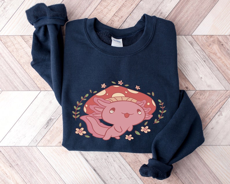 Peut inclure: Un sweat-shirt bleu marine avec un mignon dessin anim&eacute; d'axolotl portant un chapeau de champignon rouge. L'axolotl est entour&eacute; de feuilles vertes et de fleurs roses.