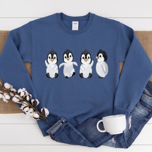 Puede incluir: Una sudadera azul con un gr&aacute;fico de cuatro ping&uuml;inos en fila. Los ping&uuml;inos son blancos con cabezas y picos negros. Todos miran hacia adelante, excepto el &uacute;ltimo ping&uuml;ino, que mira hacia la derecha.
