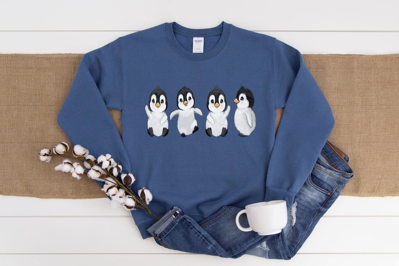 Puede incluir: Una sudadera azul con un gr&aacute;fico de cuatro ping&uuml;inos en fila. Los ping&uuml;inos son blancos con cabezas y picos negros. Todos miran hacia adelante, excepto el &uacute;ltimo ping&uuml;ino, que mira hacia la derecha.