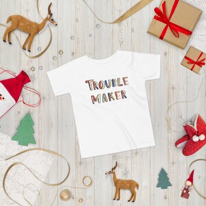 May include: White t-shirt with the words "Trouble Maker" in colorful letters. The shirt is surrounded by Christmas decorations, including a reindeer, a Christmas tree, and a gift box.
