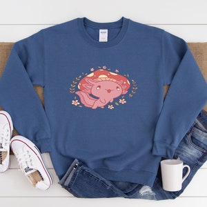 Peut inclure: Un sweat-shirt bleu avec un axolotl rose portant un chapeau de champignon rouge et entour&eacute; de fleurs.