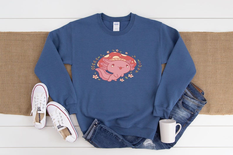 Peut inclure: Un sweat-shirt bleu avec un axolotl rose portant un chapeau de champignon rouge et entour&eacute; de fleurs.