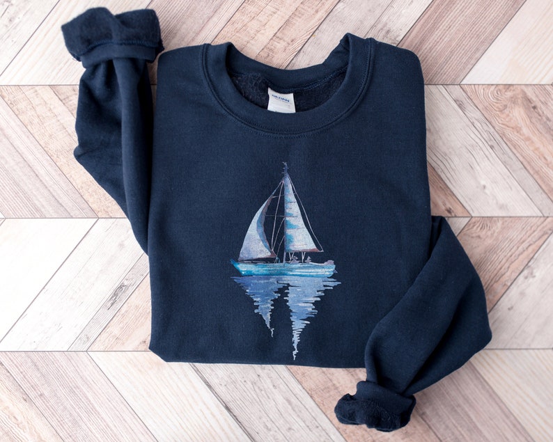 Pode incluir: Uma camisola azul marinho com um gr&aacute;fico de veleiro branco na frente. O veleiro &eacute; refletido na &aacute;gua abaixo.
