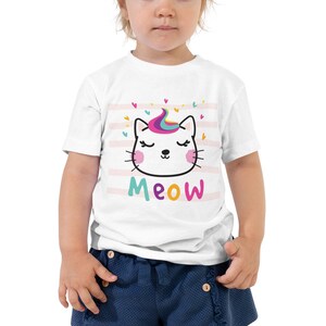 May include: White toddler t-shirt with a pink, white, and blue striped background. A white cat with a rainbow mane and pink cheeks is on the shirt. The text "MEOW" is below the cat.
