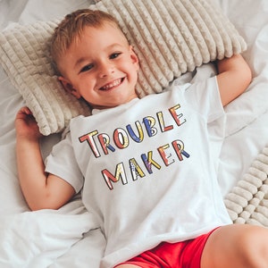 May include: A white t-shirt with the text "Trouble Maker" in colorful letters. The shirt is worn by a child lying on a white bed with a white and beige patterned blanket.