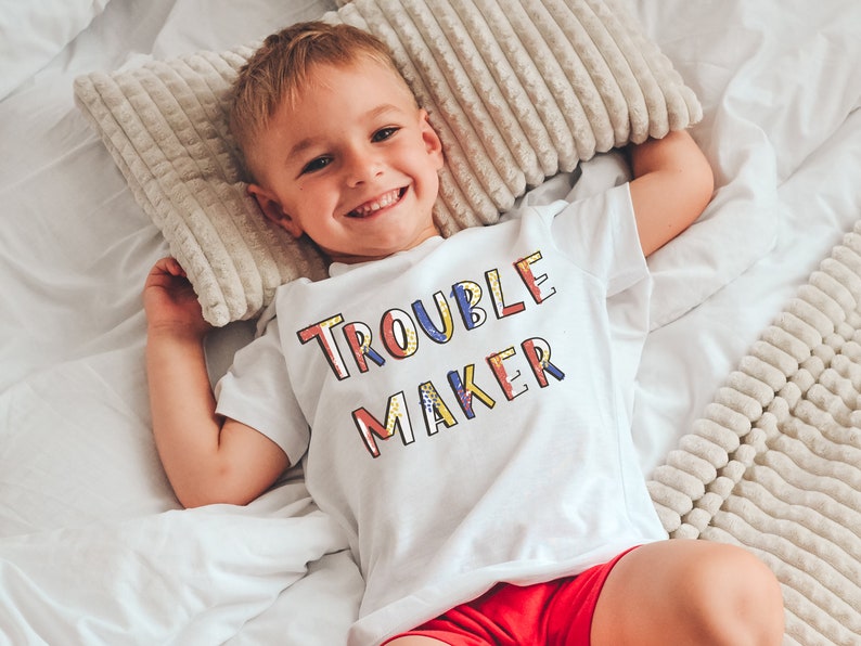 May include: A white t-shirt with the text "Trouble Maker" in colorful letters. The shirt is worn by a child lying on a white bed with a white and beige patterned blanket.