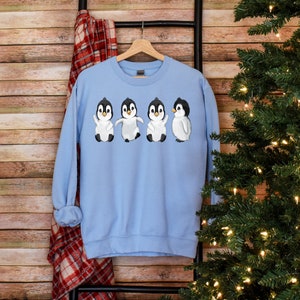 Puede incluir: Sudadera azul claro con un gr&aacute;fico de cuatro ping&uuml;inos que llevan bufandas blancas y gorros negros. Los ping&uuml;inos est&aacute;n todos mirando hacia adelante y tienen las alas extendidas.