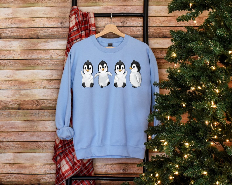 Puede incluir: Sudadera azul claro con un gr&aacute;fico de cuatro ping&uuml;inos que llevan bufandas blancas y gorros negros. Los ping&uuml;inos est&aacute;n todos mirando hacia adelante y tienen las alas extendidas.