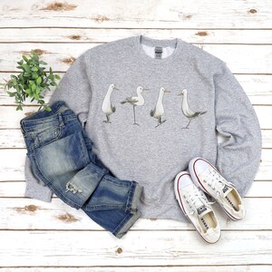 May include: A light gray sweatshirt featuring a watercolor illustration of four seagulls. The sweatshirt is paired with blue jeans and white canvas sneakers. The items are arranged on a white wooden surface.