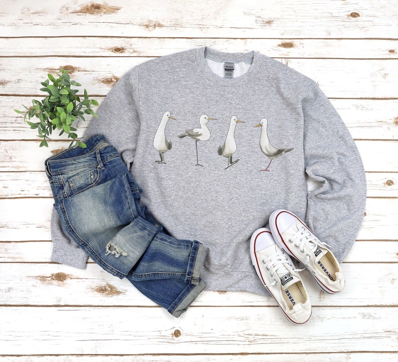 May include: A light gray sweatshirt featuring a watercolor illustration of four seagulls. The sweatshirt is paired with blue jeans and white canvas sneakers. The items are arranged on a white wooden surface.
